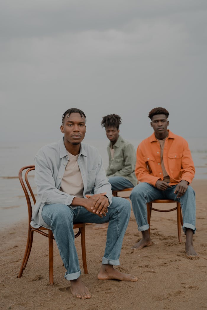 A trio of men sit barefoot on chairs along the beach under a cloudy sky.