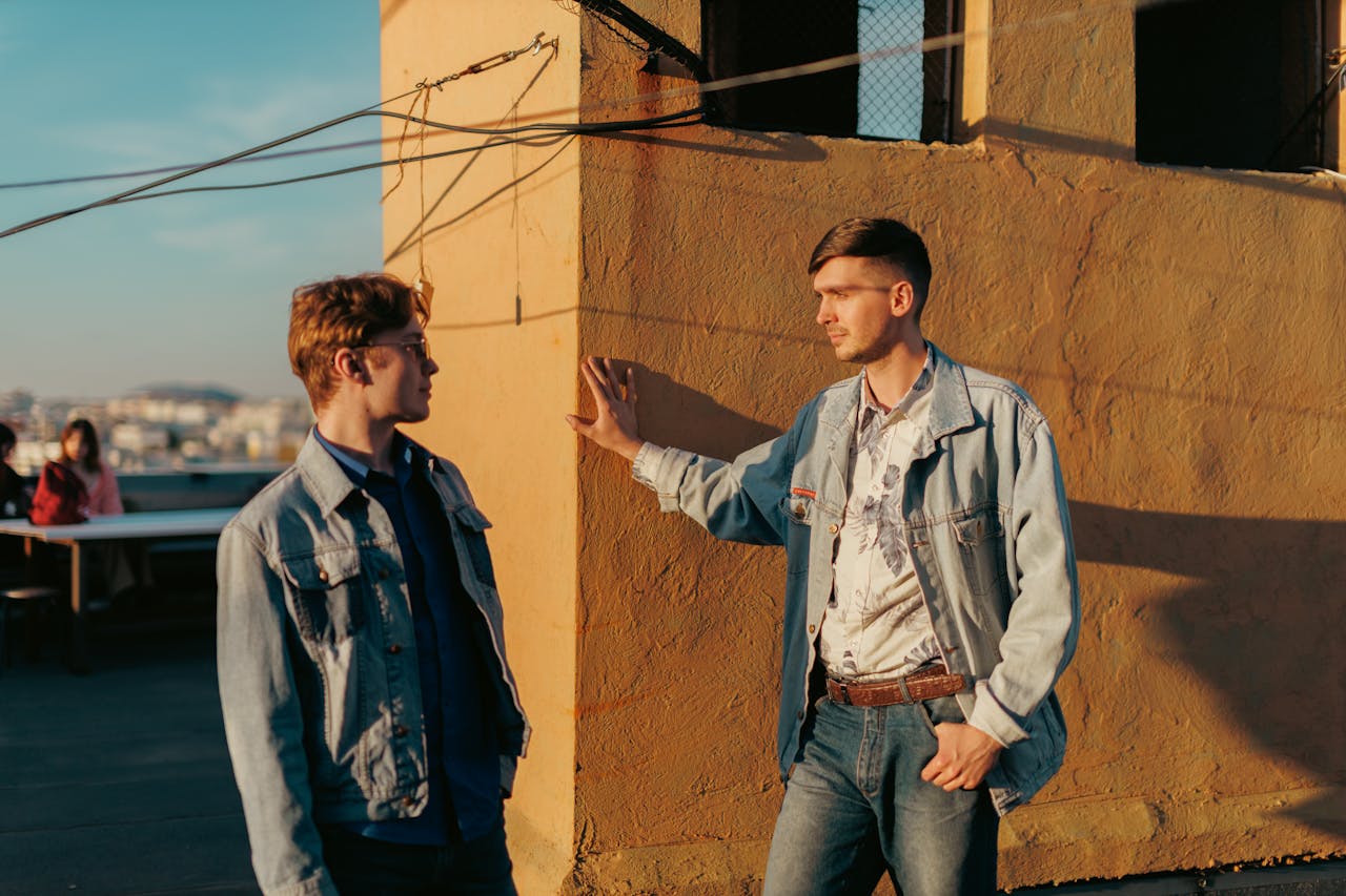 Two men wearing denim jackets engage in a casual conversation on a sunlit rooftop.