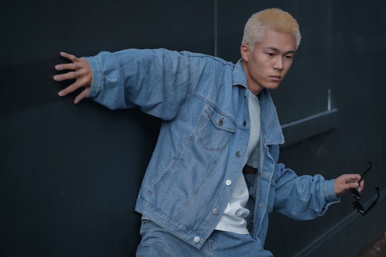Young man with dyed hair in denim jacket striking a pose with sunglasses.